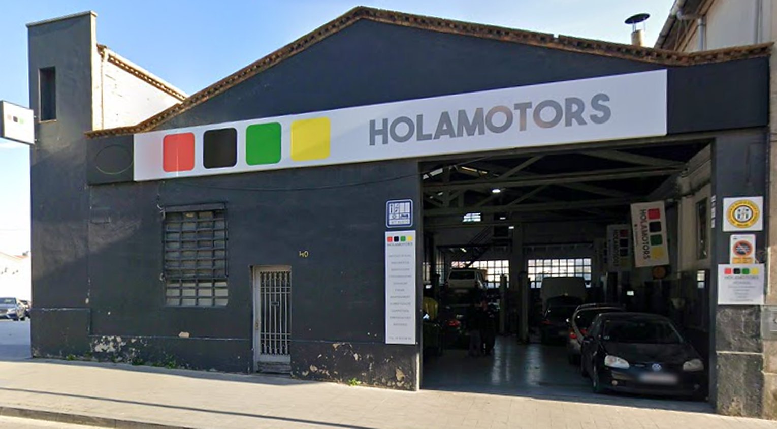 Fachada de Holamotors en la Av. de Barcelona, 40: rótulo blanco con los cuatro cuadrados de colores y el portón del taller abierto